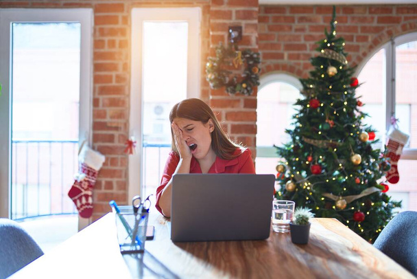 クリスマスツリーの周りの自宅でノートパソコンで作業テーブルに座って美しい女性半分の顔、目と手で口をカバー疲れている。痛みで顔が痛い. - 写真・画像
