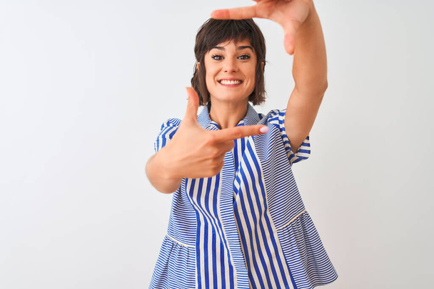 Jonge mooie vrouw draagt blauw gestreepte shirt staan over geïsoleerde witte achtergrond glimlachend maken frame met handen en vingers met gelukkig gezicht. Creativiteit en fotografie concept. - Foto, afbeelding