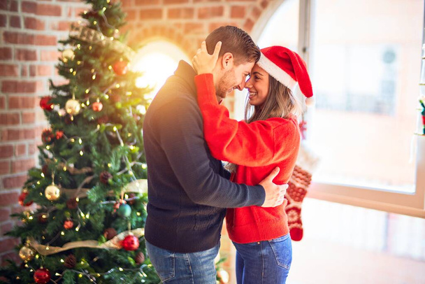 Jovem casal bonito sorrindo feliz e confiante. De pé e abraçando em torno da árvore de natal em casa
 - Foto, Imagem