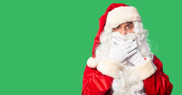 Ein gutaussehender Mann mittleren Alters im Weihnachtsmann-Kostüm und mit Bart steht schockiert da und verdeckt den Mund mit den Händen. Geheimes Konzept. - Foto, Bild
