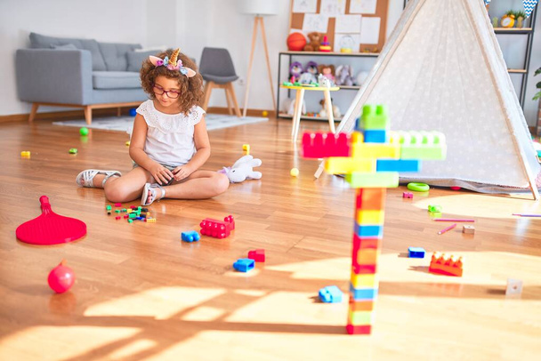 Beautiful toddler wearing glasses and unicorn diadem sitting on the floor playing with building blocks at kindergarten - Foto, immagini