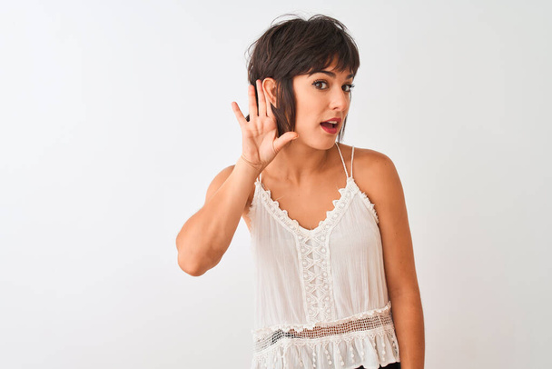 Mladá krásná žena v letním neformálním tričku stojí nad izolovaným bílým pozadím a usmívá se s rukou přes ucho a poslouchá drby nebo drby. Koncept hluchoty. - Fotografie, Obrázek