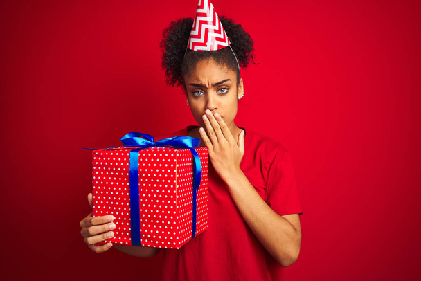 African american woman wearing funny hat holding birthday gift over isolated red background cover mouth with hand shocked with shame for mistake, expression of fear, scared in silence, secret concept - 写真・画像