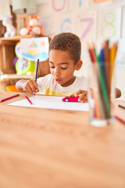 美しいアフリカ系アメリカ人の幼児は、幼稚園で机の上に紙と鉛筆を使って絵を描く - 写真・画像