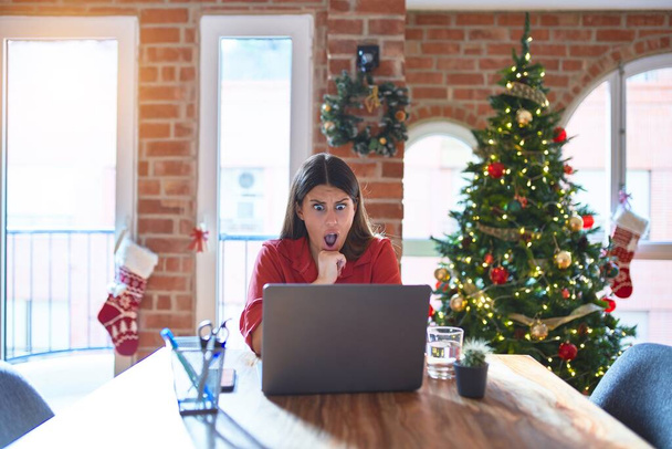 Piękna kobieta siedząca przy stole pracująca z laptopem w domu wokół choinki W szokującej twarzy, wyglądająca sceptycznie i sarkastycznie, zaskoczona otwartymi ustami - Zdjęcie, obraz