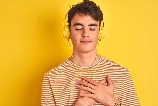 Teenager-Junge mit Kopfhörern über gelbem Hintergrund, lächelnd mit geschlossenen Augen und dankbarer Geste im Gesicht. Gesundheitskonzept. - Foto, Bild