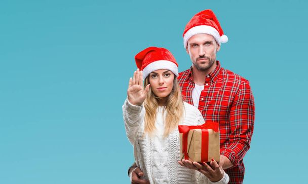 Young couple in love wearing christmas hat and holding present over isolated background with open hand doing stop sign with serious and confident expression, defense gesture - Foto, immagini