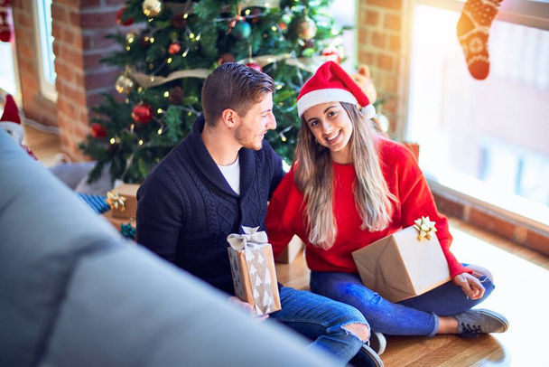 Fiatal szép pár mosolyog boldog és magabiztos. Ülni a padlón ajándékokkal a kezében és ölelkezni a karácsonyfa körül otthon. - Fotó, kép