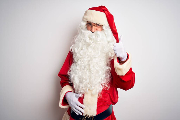 Middle age handsome man wearing Santa costume standing over isolated white background doing happy thumbs up gesture with hand. Approving expression looking at the camera showing success. - 写真・画像
