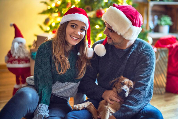 Молодая красивая пара улыбается счастливой и уверенной.. Сидя на полу в шляпе Санты Клауса, обнимая собаку вокруг рождественской елки дома
 - Фото, изображение