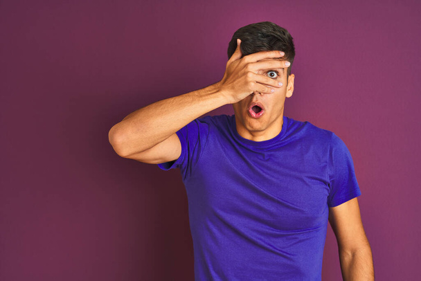 Homem indiano jovem vestindo camiseta em pé sobre fundo roxo isolado espreitando em choque cobrindo rosto e olhos com a mão, olhando através de dedos com expressão embaraçosa
. - Foto, Imagem