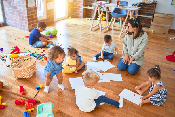 Όμορφη δασκάλα και ομάδα μικρών παιδιών που κάθονται στο πάτωμα ζωγραφίζοντας χρησιμοποιώντας χαρτί και μολύβι γύρω από πολλά παιχνίδια στο νηπιαγωγείο - Φωτογραφία, εικόνα