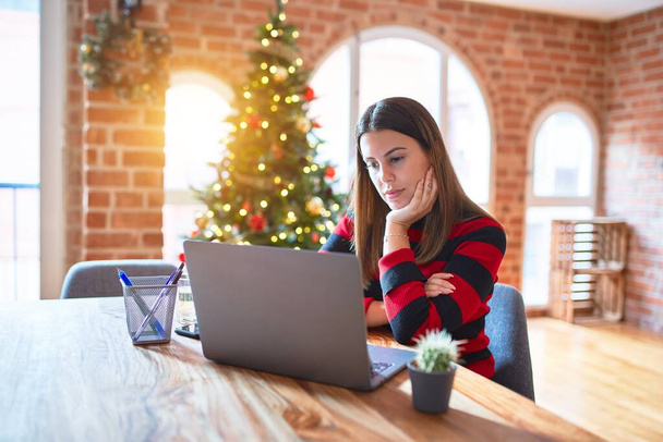 クリスマスツリーの周りの自宅でラップトップで作業テーブルに座って美しい女性が顔に深刻な表現でリラックスしました。シンプルで自然なカメラを見て. - 写真・画像