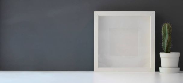 白いテーブルと暗い灰色の壁の背景にモダンなワークスペースでポットにサボテンとフレームをモックアップ - 写真・画像