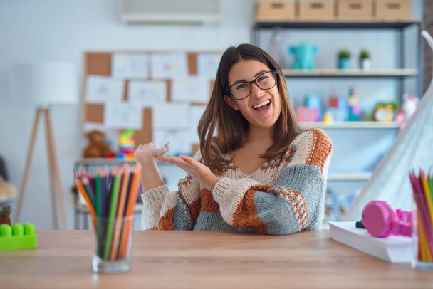 Nuori kaunis opettaja nainen yllään villapaita ja lasit istuu pöydällä päiväkodissa kutsuen sisään hymyilevä luonnollinen avoimella kädellä
 - Valokuva, kuva