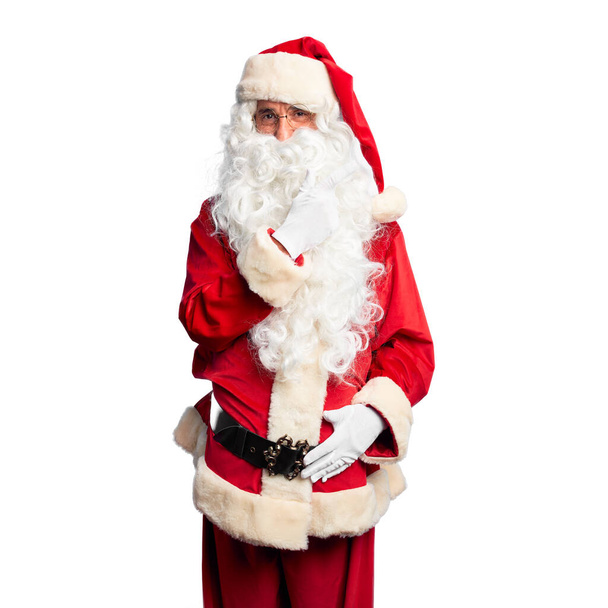 Przystojny mężczyzna w średnim wieku, ubrany w kostium Świętego Mikołaja i brodę, stojący wesoły z uśmiechem na twarzy, wskazujący ręką i palcem na bok, ze szczęśliwym i naturalnym wyrazem twarzy. - Zdjęcie, obraz