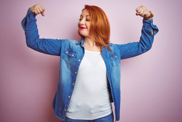 Youg beautiful redhead woman wearing denim shirt standing over isolated pink background showing arms muscles smiling proud. Fitness concept. - Фото, изображение