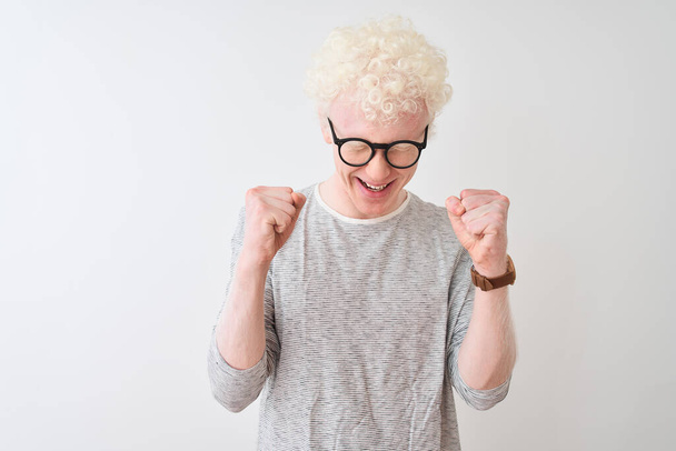 junger Albino-Blonder mit gestreiftem T-Shirt und Brille vor isoliertem weißem Hintergrund, der mit erhobenen Armen und geschlossenen Augen den Sieg lächelnd feiert. Siegerkonzept. - Foto, Bild