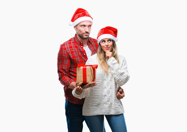 Νεαρό ζευγάρι στην αγάπη φορώντας καπέλο Χριστουγέννων και κρατώντας παρόν πέρα από το απομονωμένο φόντο σοβαρό πρόσωπο σκέψης σχετικά με την ερώτηση, πολύ συγκεχυμένη ιδέα - Φωτογραφία, εικόνα