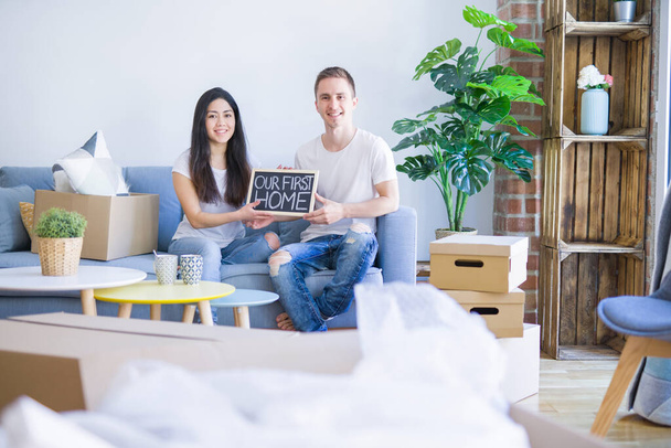 Young beautiful couple sitting on the sofa holding blackboard with message at new home around cardboard boxes - Photo, Image