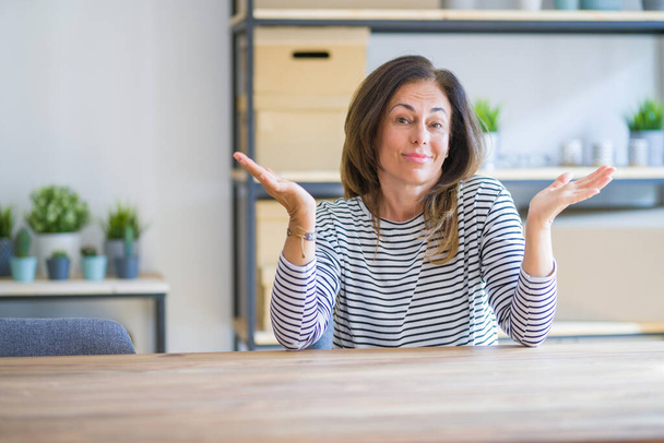 Middle age senior woman sitting at the table at home clueless and confused expression with arms and hands raised. Doubt concept. - Fotoğraf, Görsel