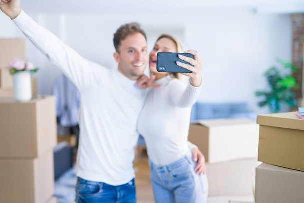 段ボール箱の周りに新しい家で自分撮りを取るためにスマートフォンを使用して立っている若い美しいカップル - 写真・画像