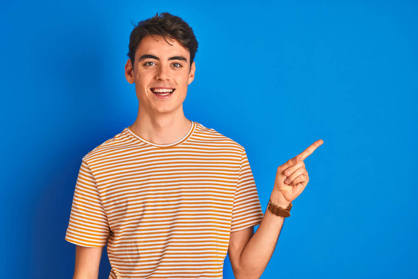 Tinédzser fiú alkalmi pólót visel, kék elszigetelt háttér felett áll egy nagy mosollyal az arcán, mutató kézzel és ujjal oldalra nézi a kamerát. - Fotó, kép