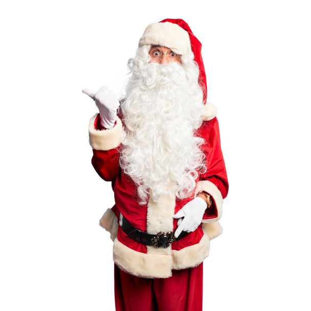 Middle age handsome man wearing Santa Claus costume and beard standing Surprised pointing with hand finger to the side, open mouth amazed expression. - Photo, Image