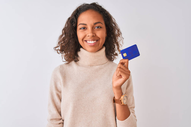 Молода бразіліанка, яка тримає кредитну картку, стоїть на ізольованому білому фоні з щасливим обличчям і усміхається з упевненою усмішкою, показуючи зуби. - Фото, зображення