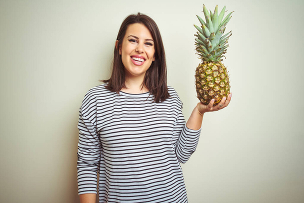 Fiatal gyönyörű nő tartja trópusi gyümölcs ananász felett elszigetelt háttér egy boldog arc áll, és mosolyog magabiztos mosollyal mutatja fogak - Fotó, kép