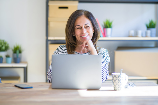 Middle age senior woman sitting at the table at home working using computer laptop looking confident at the camera smiling with crossed arms and hand raised on chin. Thinking positive. - 写真・画像