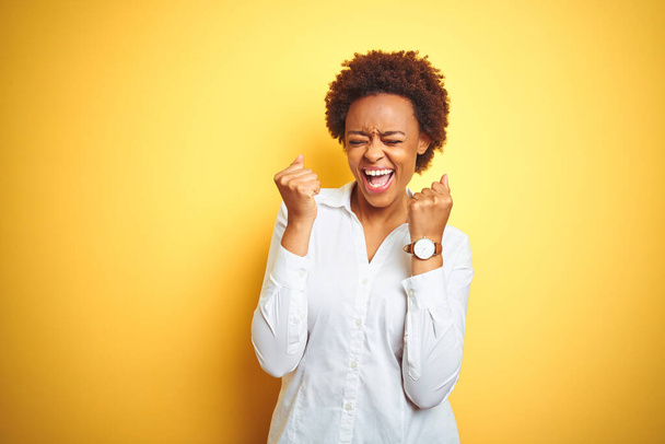 Afrykańska amerykańska kobieta biznesu na odizolowanym żółtym tle podekscytowana sukcesem z podniesionymi ramionami i zamkniętymi oczami świętującymi zwycięstwo uśmiechając się. Koncepcja zwycięzcy. - Zdjęcie, obraz