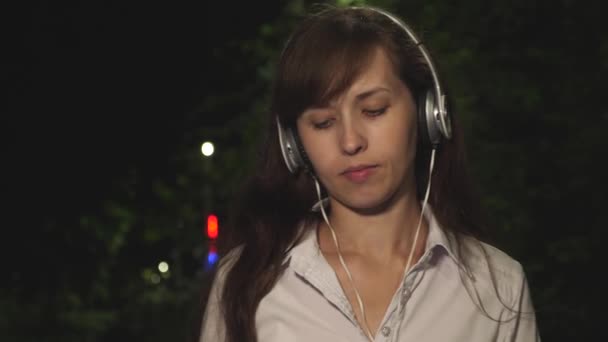 Chica en auriculares incluye canción en el teléfono táctil agitando su pelo largo y bailando sonriendo y kayfuya de la música por la noche en el parque de la ciudad. Primer plano
 - Metraje, vídeo