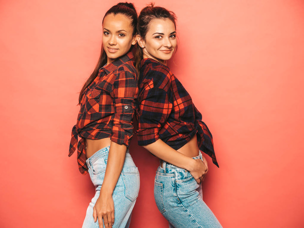 Two young beautiful smiling brunette hipster girls in trendy similar checkered shirt and jeans clothes.Sexy carefree women posing near pink wall in studio.Positive models having fun - Foto, Bild