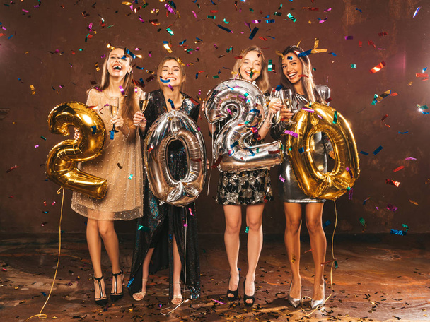 Belles femmes célébrant le Nouvel An. Filles magnifiques heureux dans des robes de soirée sexy élégantes tenant or et argent 2020 ballons, s'amuser à la fête du Nouvel An. Arrangeant et buvant des flûtes à champagne
 - Photo, image