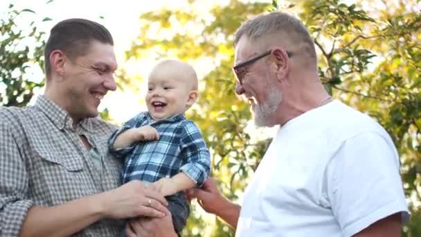 Dziadek, syn i wnuk śmieją się wesoło na tle promieni zachodzącego słońca. Szczęśliwa rodzina bawi się w sadach jabłkowych. Dzień ojców - Materiał filmowy, wideo