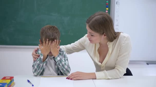 kind teacher calms crying scholar boy during extracurricular activities at desk near blackboard in classroom of school - Filmagem, Vídeo
