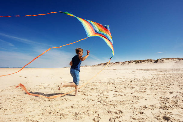 Chłopiec biegać na plaży piasku gospodarstwa kolorowe tęczy pozbawione latawca widok z tyłu - Zdjęcie, obraz