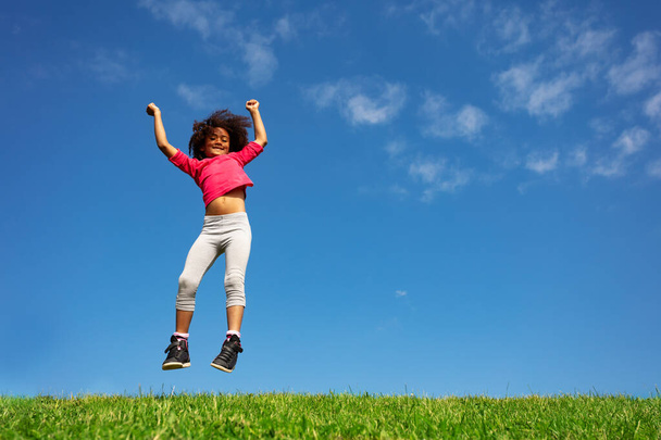 Happy little girl with curly hair jump over blue sky and clouds on the grass field hands lifted smiling - Photo, Image