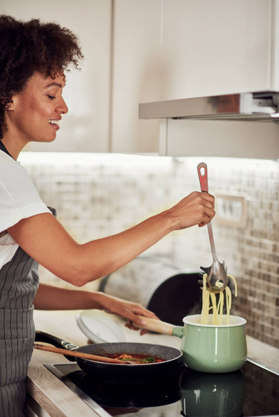 Beautiful mixed race woman in apron standing next to stove and making spaghetti. - Foto, Bild