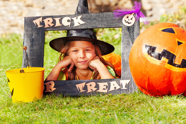 Beautiful little girl in Halloween costume look through frame with trick or treat sign near pumpkin - Foto, Imagem