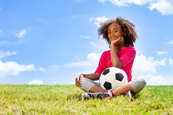 Beautiful black girl sit on the grass with football ball and putting palm to chin - Photo, image