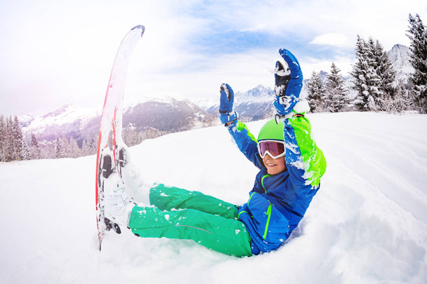 スキーウェアを着た幸せな少年は雪の中で手を持ち上げ、背景の山の上で笑顔を楽しんでいます - 写真・画像