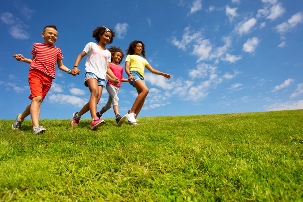 Группа разноликих детей бежит по траве, держа руки над чистым голубым небом.
 - Фото, изображение