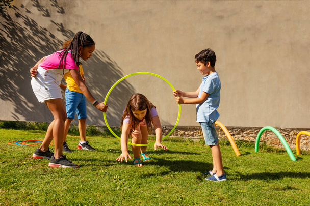 Group of friends play with color sport hoops, little long hair girl jumps inside hold by two kids - Foto, afbeelding
