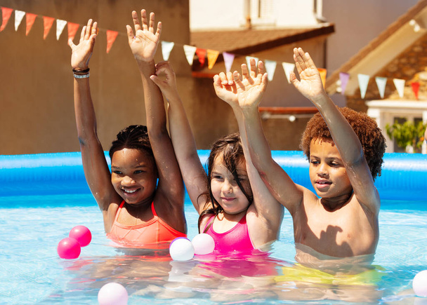 Group of four happy kids boys and girls play in the backyard swimming pool on hot summer day lifting hands up smiling - 写真・画像