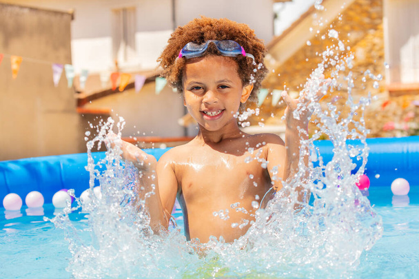 Happy handsome black boy with curly hair and wearing sport googles splash in swimming pool - Zdjęcie, obraz