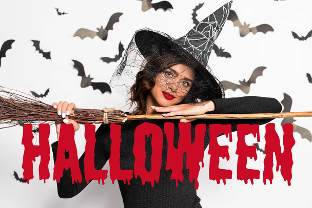 attractive woman in witch hat holding broom in Halloween near Halloween red lettering - Photo, Image