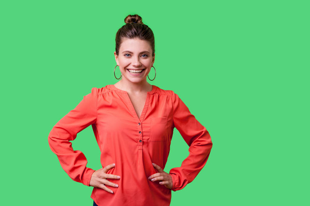 confident positive young woman with bun hairstyle and big earrings in red blouse holding hands on hips and lovely smiling at camera on green background - Photo, Image