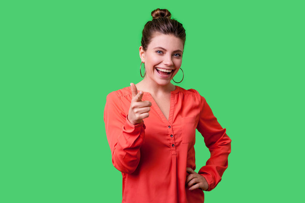 lovely positive woman with bun hairstyle and big earrings in red blouse happily pointing finger at camera and smiling on green background - Foto, Imagen